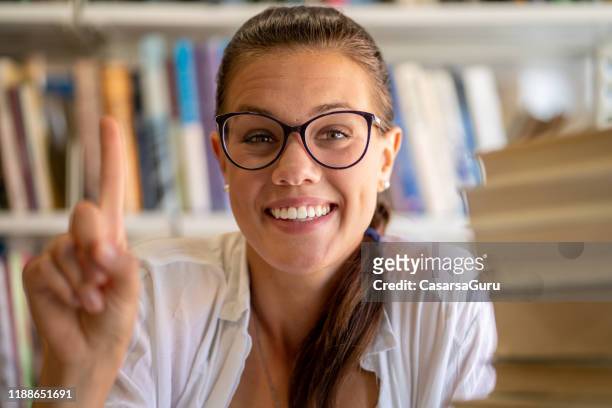 verticale de belle jeune femme adulte de sourire avec le doigt soulevé dans la bibliothèque - doigt levé photos et images de collection
