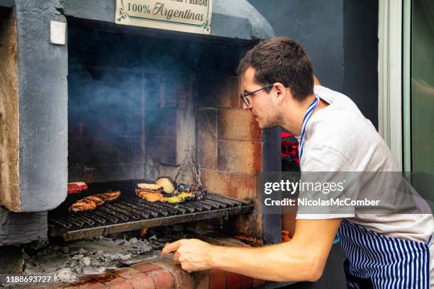 joven chef argentino agitando brasas en su parrilla de parilla mientras sus carnes cocinan - argentina steak fotografías e imágenes de stock