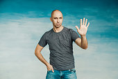 Attractive bald man on a blue background raised his left hand up with his  palm up for greetings,  towards the camera