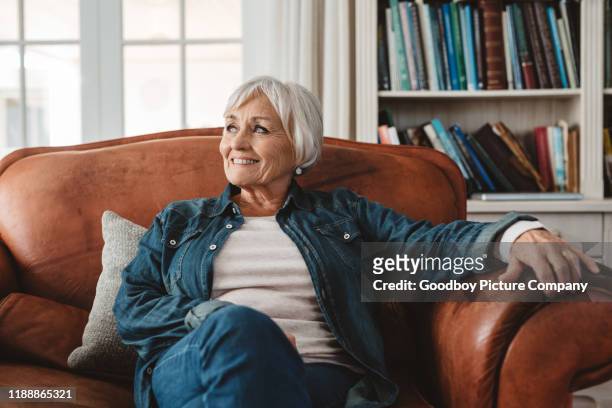 sonriendo mujer mayor sentada en su sofá en casa - comfortable fotografías e imágenes de stock