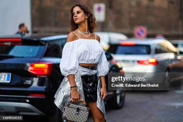 Guest wears earrings, necklaces, a white strappy off-the-shoulder gathered crop top with puff sleeves, a shiny silver-tone jacket around the waist, a...