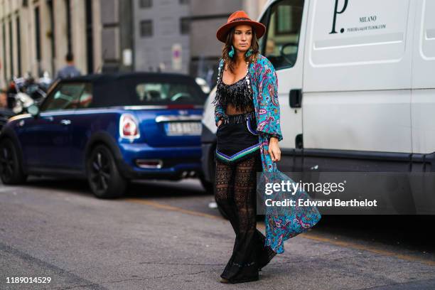 Guest wears a rust-color hat, green earrings, a glittering black plumetis top with iridescent beadwork, a black fringed mini skirt trimmed with blue,...