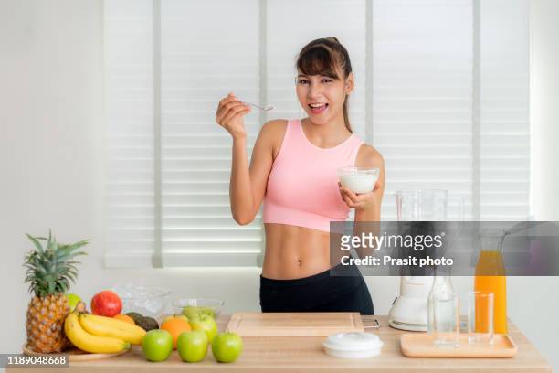 asian young woman wearing pink sportware holding yogurt for squeezing mix friut juice smoothie with apple,orange, banana, pineapple and avocado at kitchen table. healthy food and diet lifestyle for slim and good eating concept - woman squeezing orange stockfoto's en -beelden