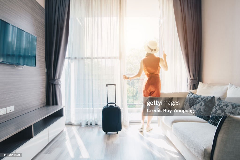Portrait of tourist woman standing nearly window, looking to beautiful view with her luggage in hotel living room after check-in.