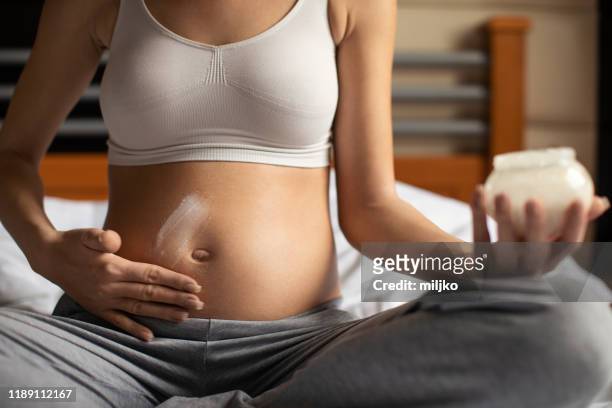 pregnant woman in bedroom applying moisturizer on her abdomen - indulgence stock pictures, royalty-free photos & images