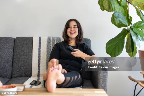 woman sitting on couch with feet up holding tv remote - businesswoman barefoot stock-fotos und bilder