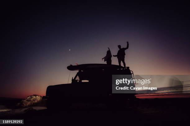 couple standing on top of a 4x4 while camping during the blue hour - couple on the beach with car stockfoto's en -beelden