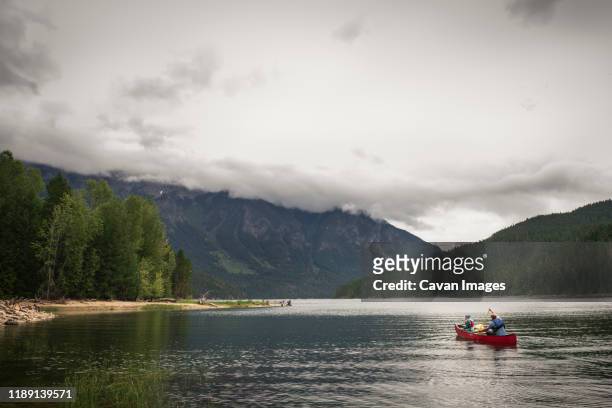 man and child paddle a red canoe on a mountain lake on a cloudy day - hiking mature man stock pictures, royalty-free photos & images