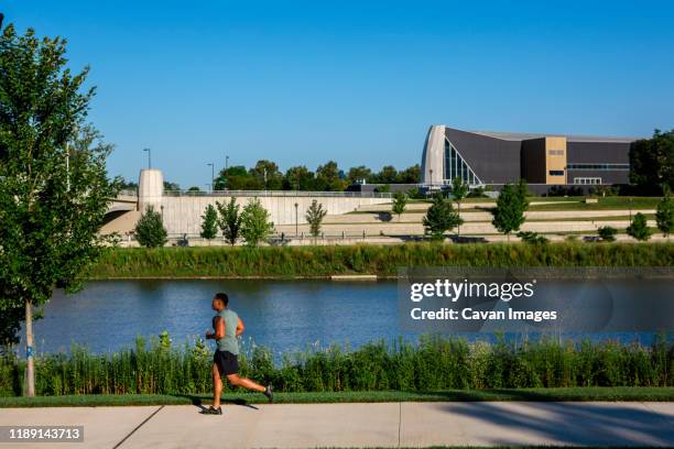 a man jogs along path in a city park against river and tall buildings - columbus ohio neighborhood stock pictures, royalty-free photos & images