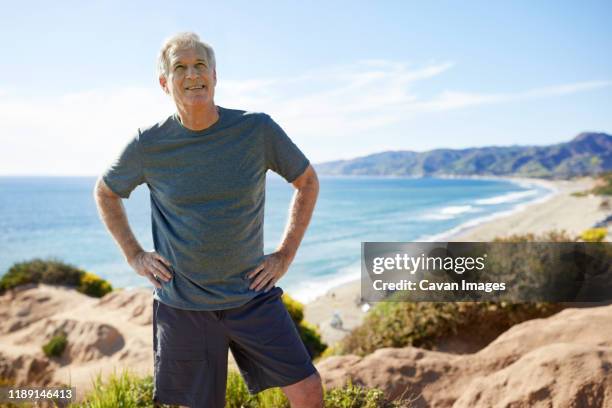 smiling senior man with hands on hips standing on cliff at beach against sky - hand on hip stock pictures, royalty-free photos & images