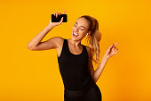 Woman In Wireless Earbuds Holding Smartphone And Dancing, Studio Shot