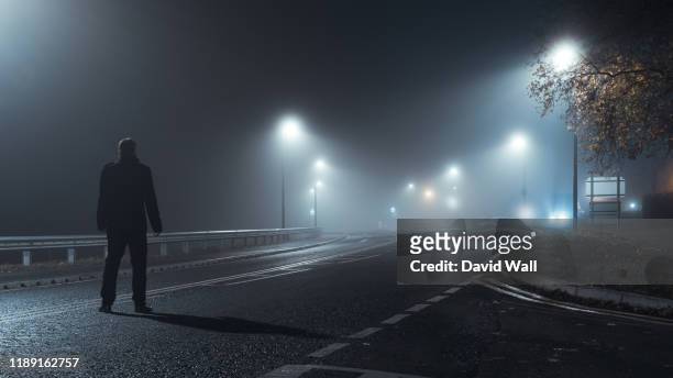 a man standing to his back to the camera, wearing a winter coat. looking down an empty road on a moody, atmospheric, spooky night. on a foggy winters evening. - dark country road stock pictures, royalty-free photos & images