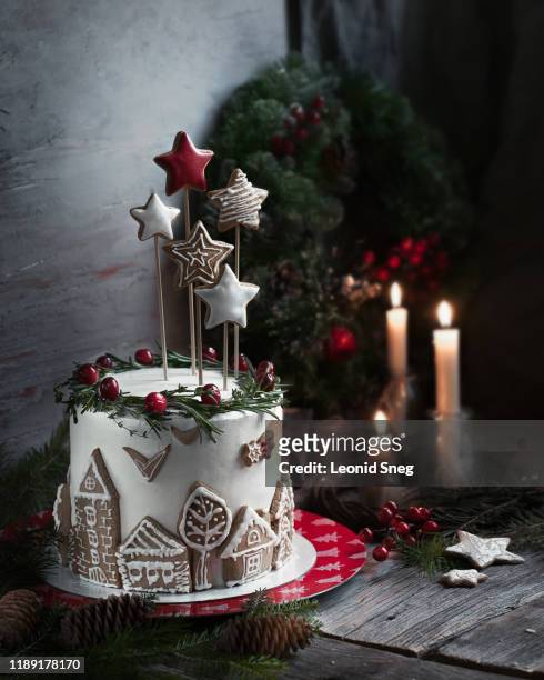 christmas white cream cake with star decor and gingerbread houses with green spicy branches and red berries on a red and white dish on a background of a fir wreath and burning candles - christmas cake foto e immagini stock