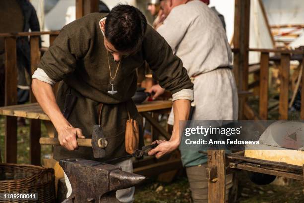 reconstruction of old crafts. a craftsman in historical clothing is hammering on the anvil. a blacksmith forges a metal product. dressed in an old outfit. - historical reenactment foto e immagini stock