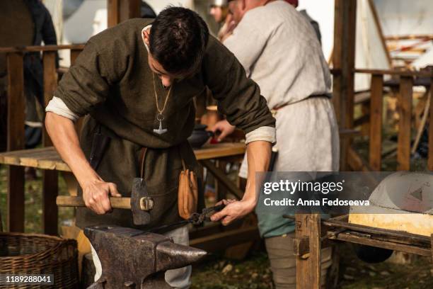 reconstruction of old crafts. a craftsman in historical clothing is hammering on the anvil. a blacksmith forges a metal product. dressed in an old outfit. - reconstituição - fotografias e filmes do acervo