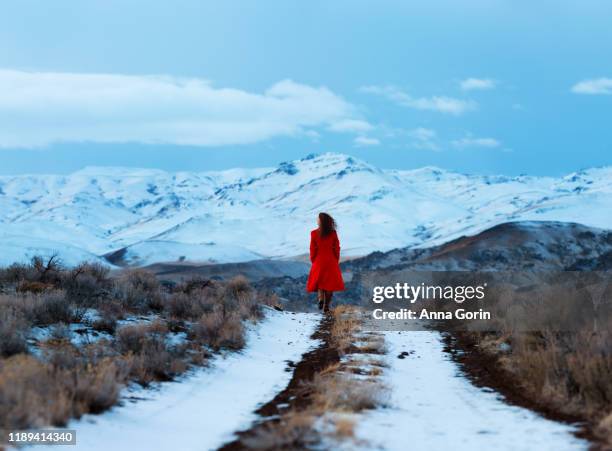 back view of young woman in long red coat walking down snow-covered two-track dirt road toward snow-covered mountains - casaco comprido vermelho imagens e fotografias de stock