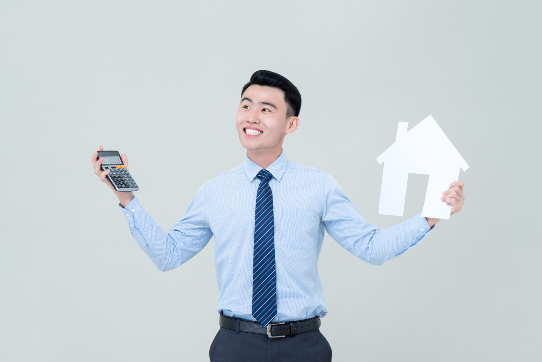 Young smiling Asian male real estate agent holding calculator and house cutout Young smiling Asian male real estate agent holding calculator and house cutout