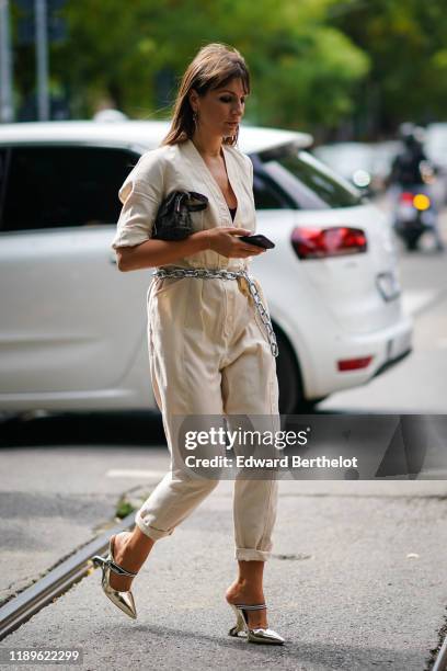 Carlotta Rubaltelli wears earrings, a necklace, a Fendi bag, a cream-color jumpsuit with a plunging neckline and rolled-up hem pants, a chain belt...