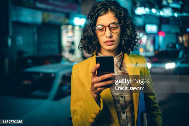 junge erwachsene geschäftsfrau liest ein smartphone auf einer belebten straße in der nacht. - india phone professional stock-fotos und bilder