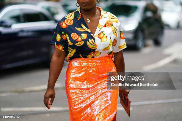 Guest wears earrings, necklaces, a half-white half-black shirt with a yellow, orange and green fruit pattern, an orange glittering sequined side-slit...