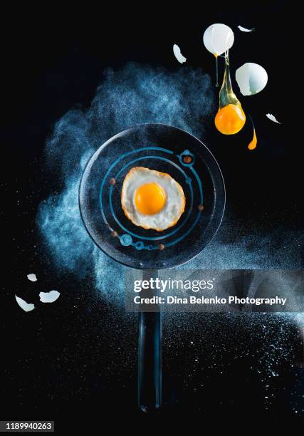 frying pen with en egg in a cloud of flour and falling yolk on a black bachground. space breakfast concept - broken plate stock pictures, royalty-free photos & images