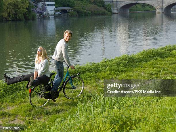 couple double on bicycle by river's edge - rivier gras oever stockfoto's en -beelden