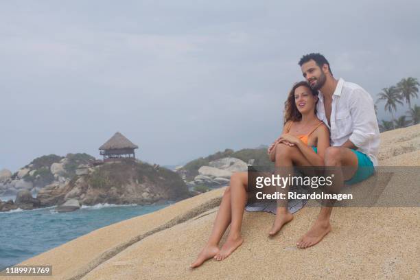 couples ethniques latins s'asseyant ensemble souriant au-dessus d'un rocher, regardant l'horizon embrassant l'un l'autre. en arrière-plan, vous pouvez voir la mer avec une forte houle, et une cabine typique de tayrona park - can beach sun photos et images de collection