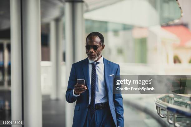 businessman walking and looking at cell phone - black suit sunglasses stock pictures, royalty-free photos & images