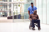 Male Nurse Wearing Scrubs Wheeling Patient In Wheelchair Through Lobby Of Modern Hospital Building