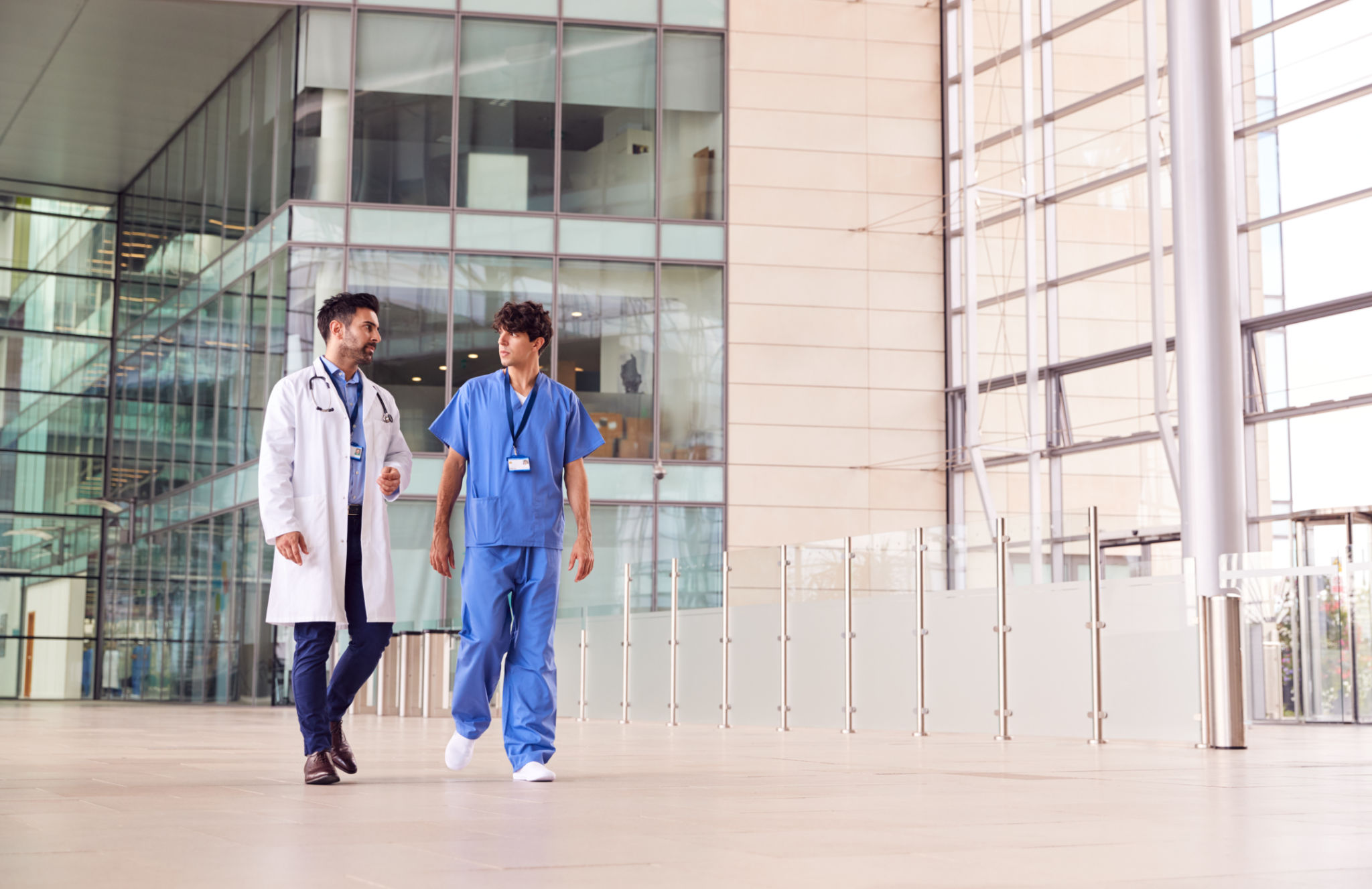 Male Medical Staff Talking As They Walk Through Lobby Of Modern Hospital Building Male Medical Staff Talking As They Walk Through Lobby Of Modern Hospital Building