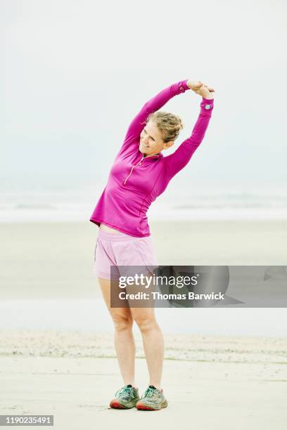 smiling mature woman stretching on beach before early morning run - knallrosa stock-fotos und bilder