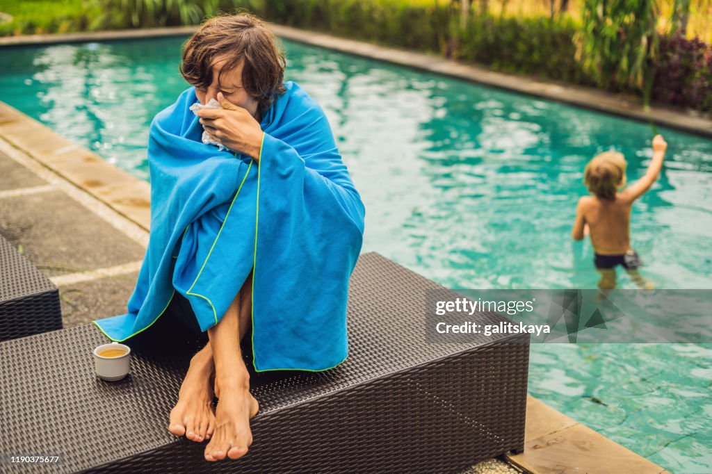 Sick man traveler. The man caught a cold on vacation, sits sad at the pool drinking tea and blows his nose into a napkin. His son is healthy and swimming in the pool. Travel insurance concept