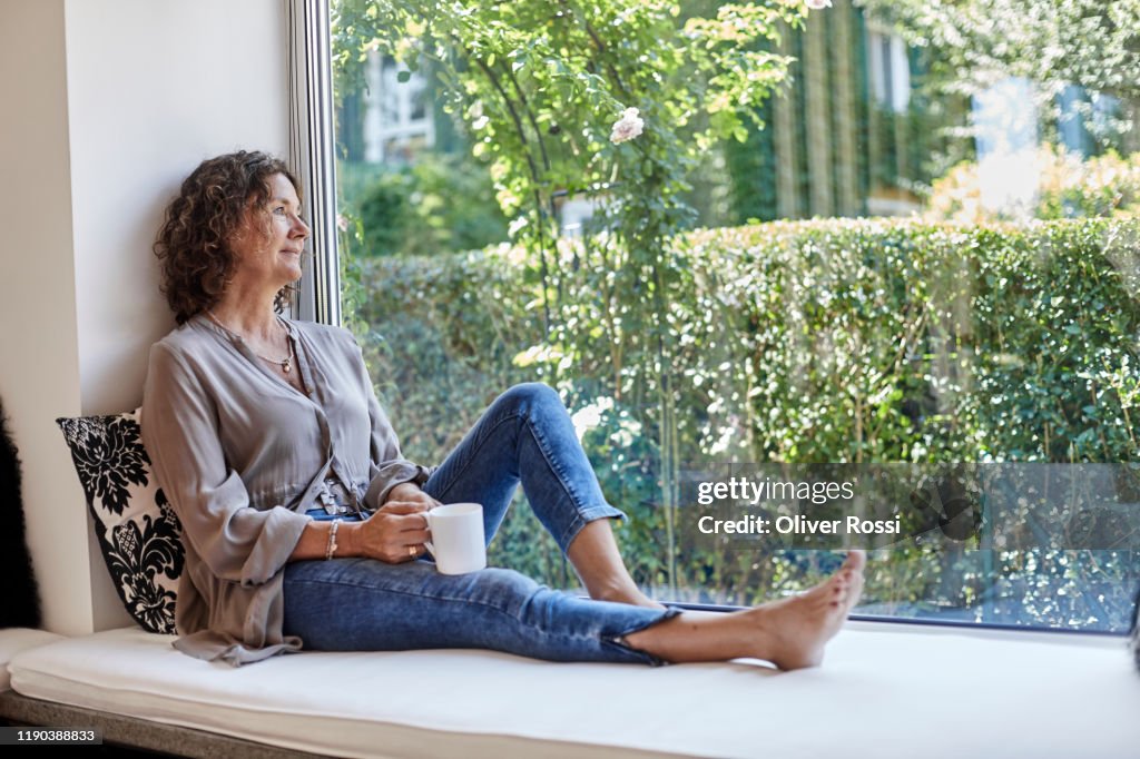 Mature woman sitting at the window at home with cup of coffee