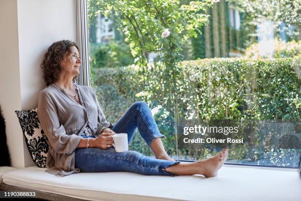 mature woman sitting at the window at home with cup of coffee - woman looking out of window photos et images de collection