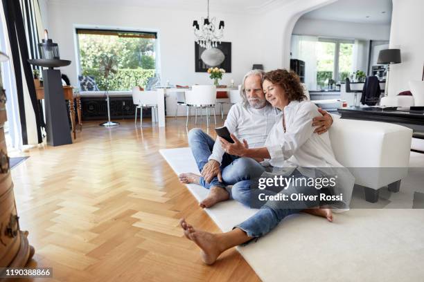 senior couple sitting on the floor at home using smartphone - auf dem boden sitzen stock-fotos und bilder