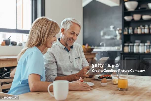 lachende volwassen paar met behulp van een digitale tablet tijdens het eten van ontbijt - couple at table with ipad stockfoto's en -beelden