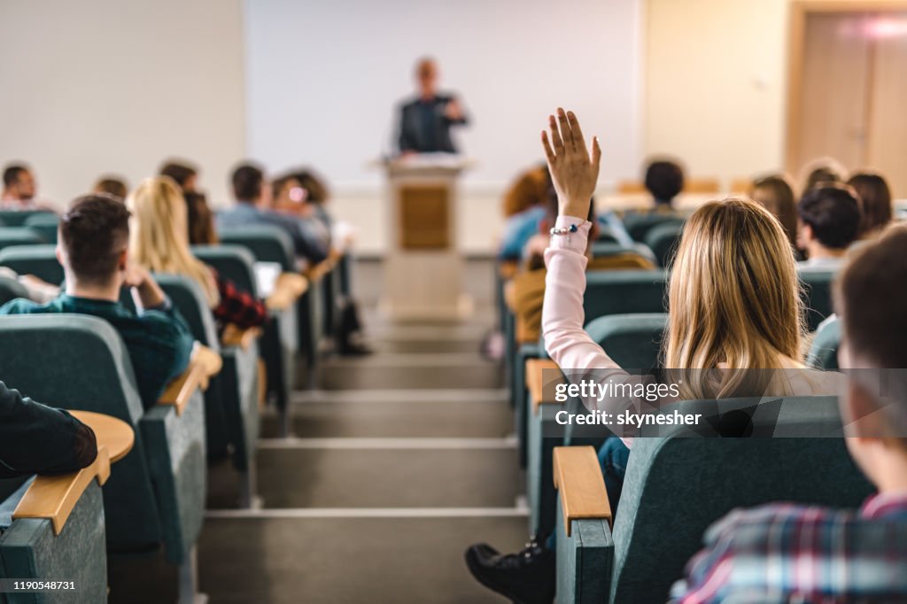 Rear view of college student raising her hand in amphitheater.