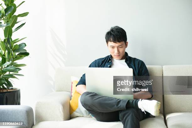 young asian man using computer in living room - asian man home laptop stock pictures, royalty-free photos & images