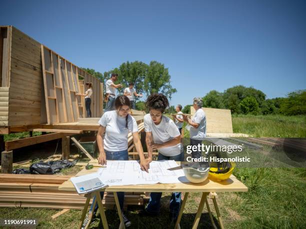 female volunteers checking the blueprint of a housing project while the team is working at background - habitat for humanity stock pictures, royalty-free photos & images