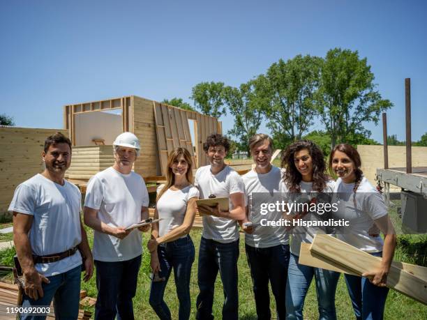 happy group of volunteers at a construction site facing camera smiling while holding tools, plywood and clipboards - habitat for humanity stock pictures, royalty-free photos & images