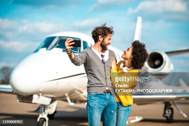 rich young couple taking a selfie on a mobile phone in front of a private jet parked on an airport tarmac - millionnaire stock pictures, royalty-free photos & images