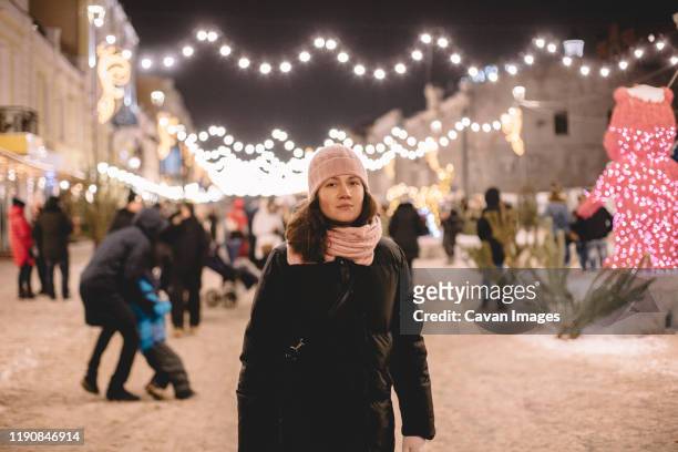 young woman wearing warm clothing standing in brightly lit street - alone in a crowd sad stockfoto's en -beelden