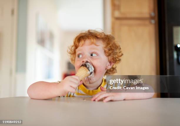 messy toddler boy eats ice cream while sitting at counter in kitchen - boy eating stock-fotos und bilder