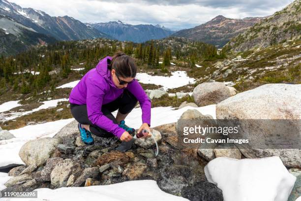 a women crouches down and uses a water filter to get drinking water from a small alpine stream in the mountains of british columbia. - hurley stick stock pictures, royalty-free photos & images
