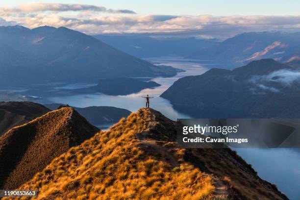 man standing at peak of mountain overlooking blue lake during sunset. - wanaka stock-fotos und bilder