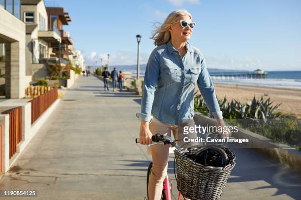 cheerful senior woman riding bicycle on road against sky in city - manhattan beach stockfoto's en -beelden