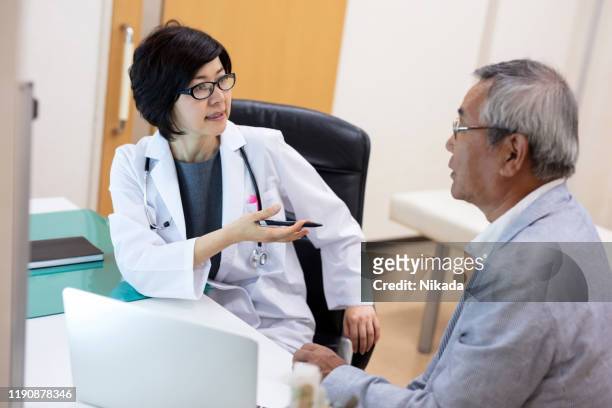 doutor fêmea que explica o paciente masculino sênior ao sentar-se na mesa no hospital - sala de exame médico - fotografias e filmes do acervo