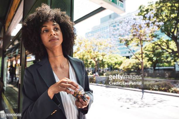 business afro amerikanische frau an bushaltestelle in der innenstadt - snacking on the go stock-fotos und bilder