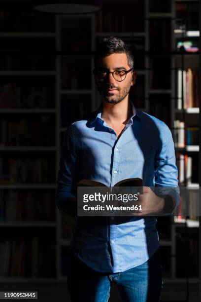 portrait of young man with book standing in front of bookshelves at home - kontrastreich stock-fotos und bilder