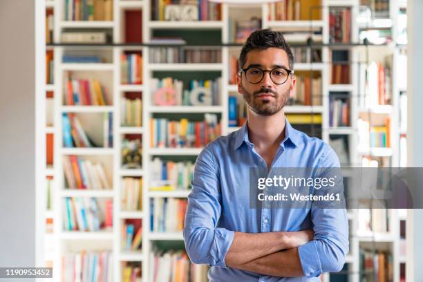 portrait of bearded young man standing in front of bookshelves at home - mezzo busto foto e immagini stock